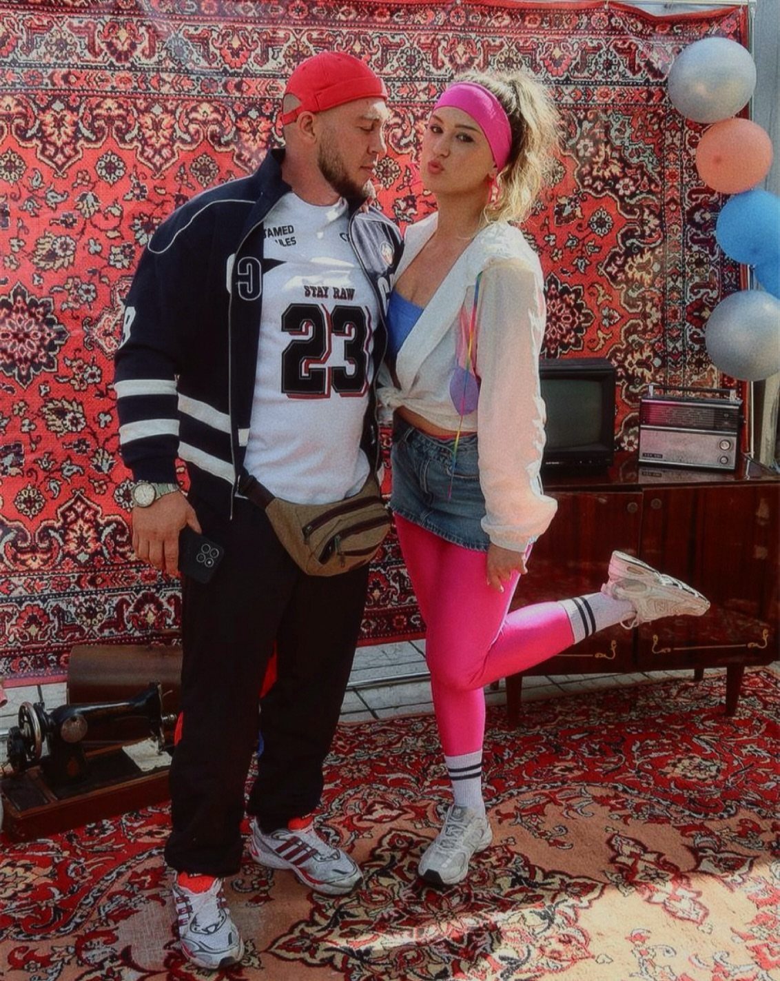 A man and woman posing in colorful 90s style outfits against a patterned rug.