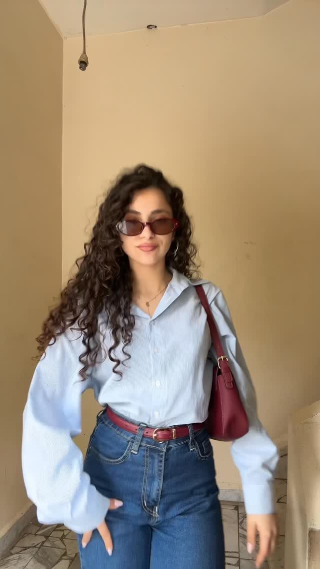 Woman with curly hair wearing rectangular sunglasses, a blue shirt, and high-waisted jeans.