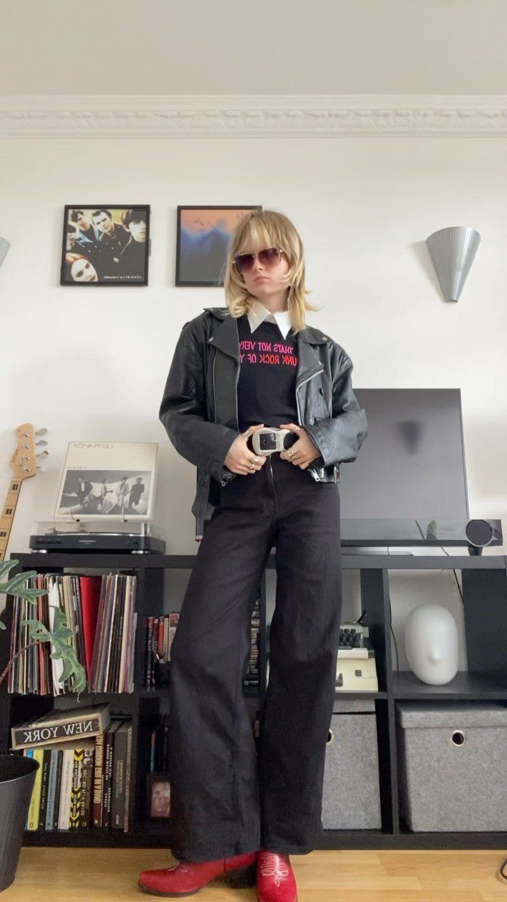 A person wearing a black leather jacket, graphic t-shirt, and black wide-leg trousers standing indoors.