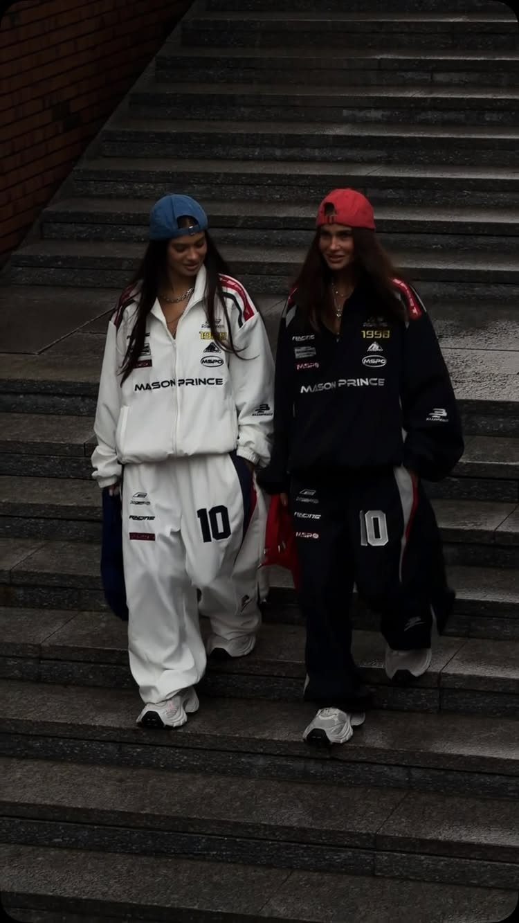 Two women walking down stairs wearing oversized athletic tracksuits and baseball caps.