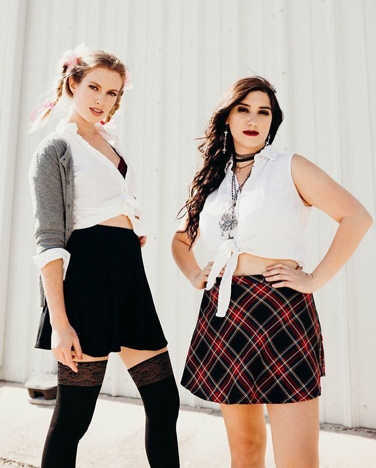 Two women posing in white tied shirts and skirts against a white wall.