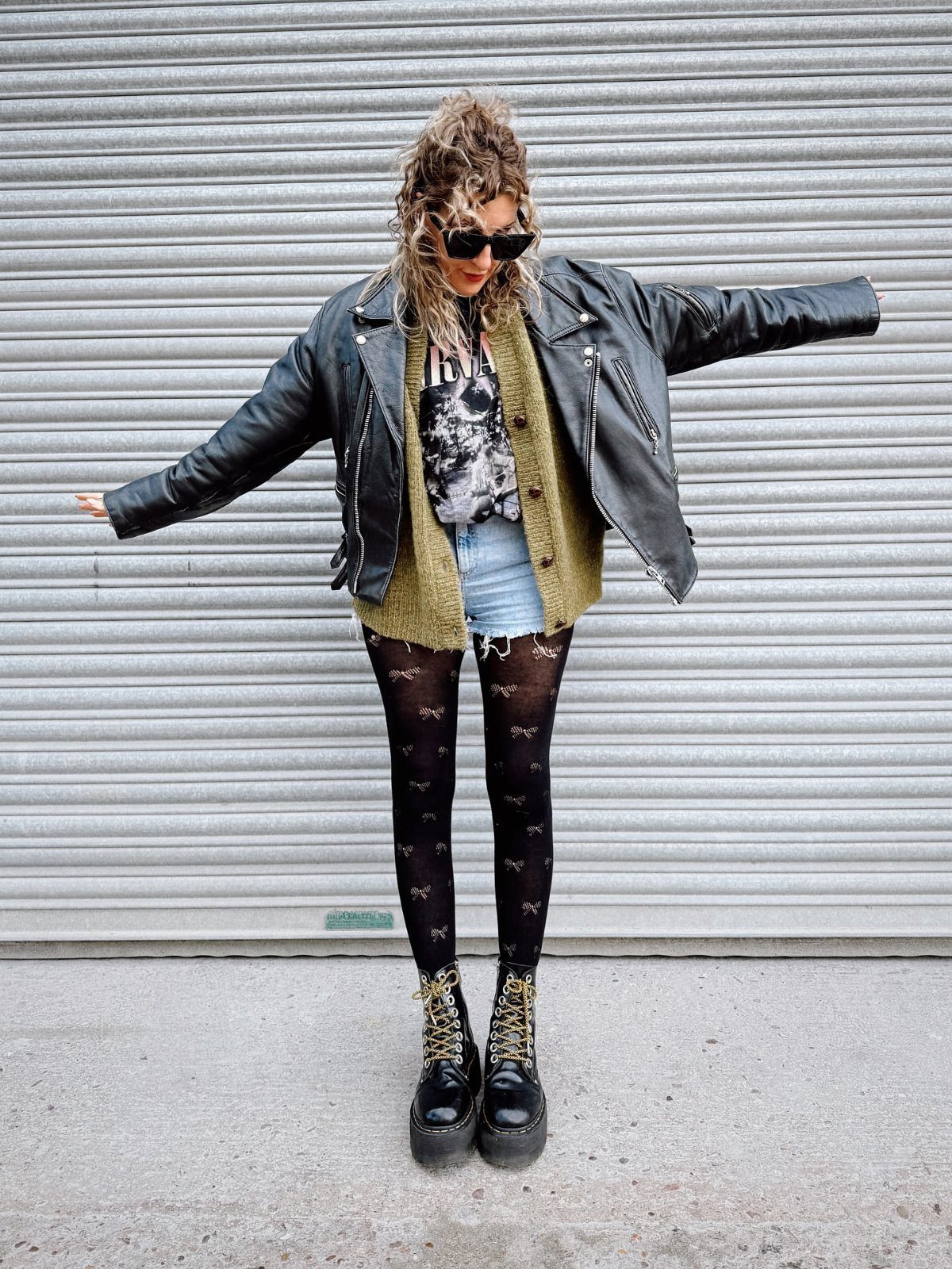 A person posing in a leather jacket, graphic tee, denim shorts, patterned tights, and combat boots.