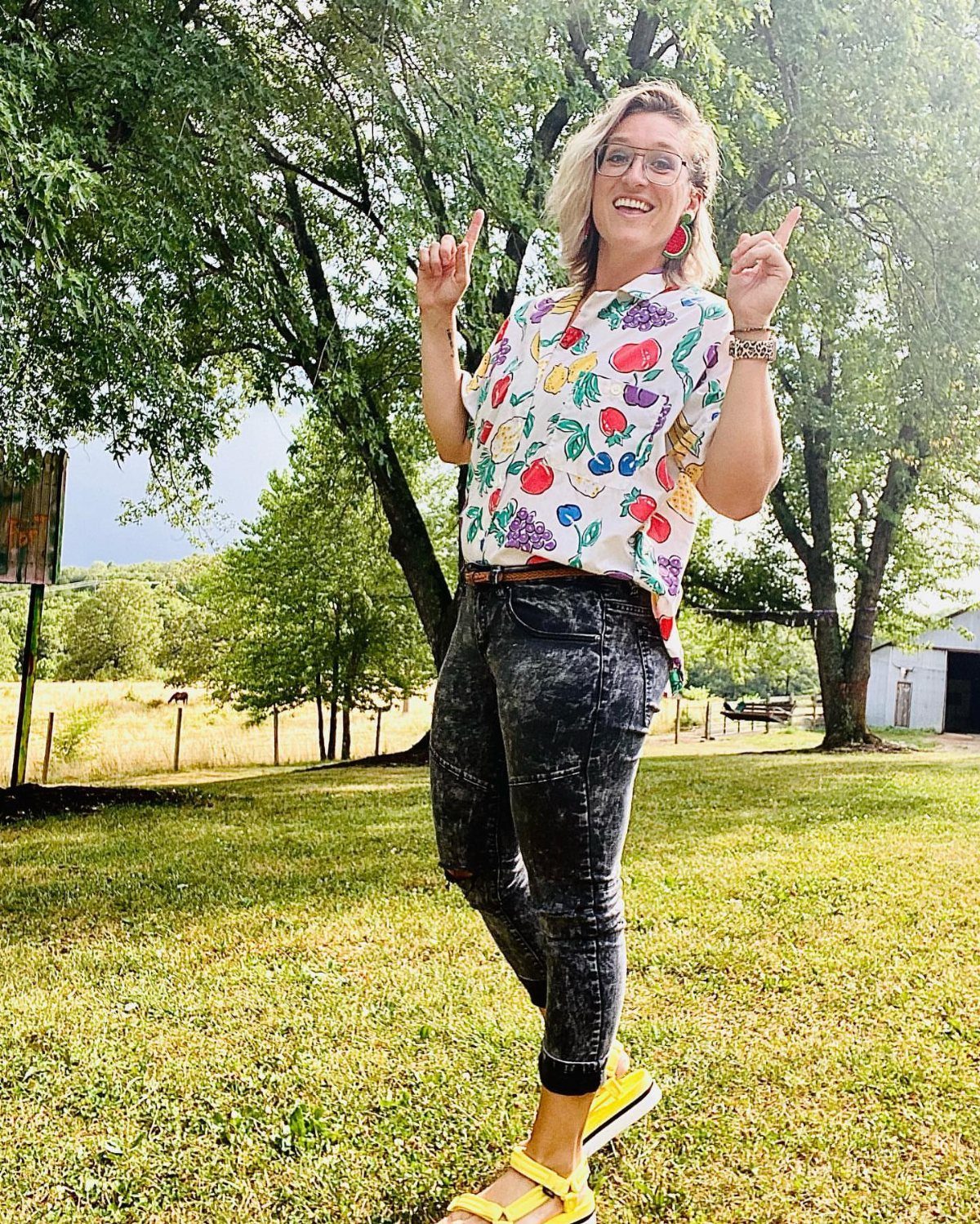 Smiling woman wearing a fruit print shirt and acid-wash jeans standing in a grassy field.