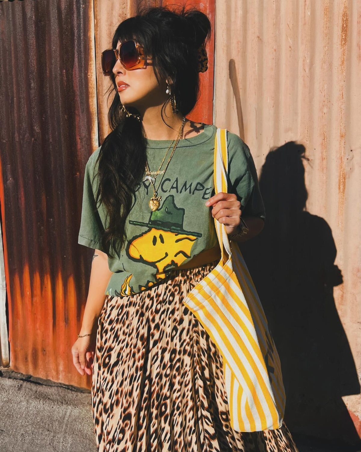 Woman in a green graphic tee and leopard print skirt standing by a metal wall.