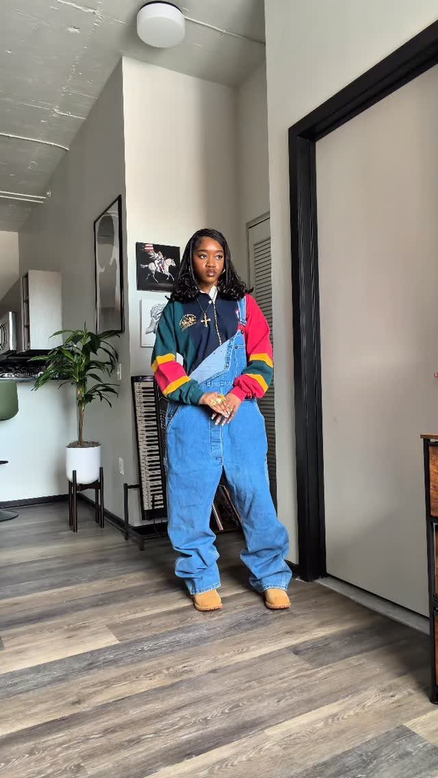 A woman posing in blue denim overalls and a colorful striped sweatshirt in a room.