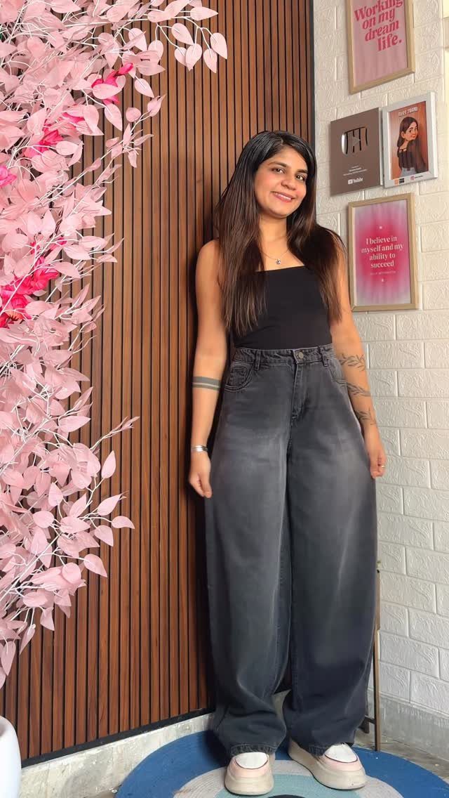 A woman posing in a black tank top and dark grey high-waisted wide-leg jeans.