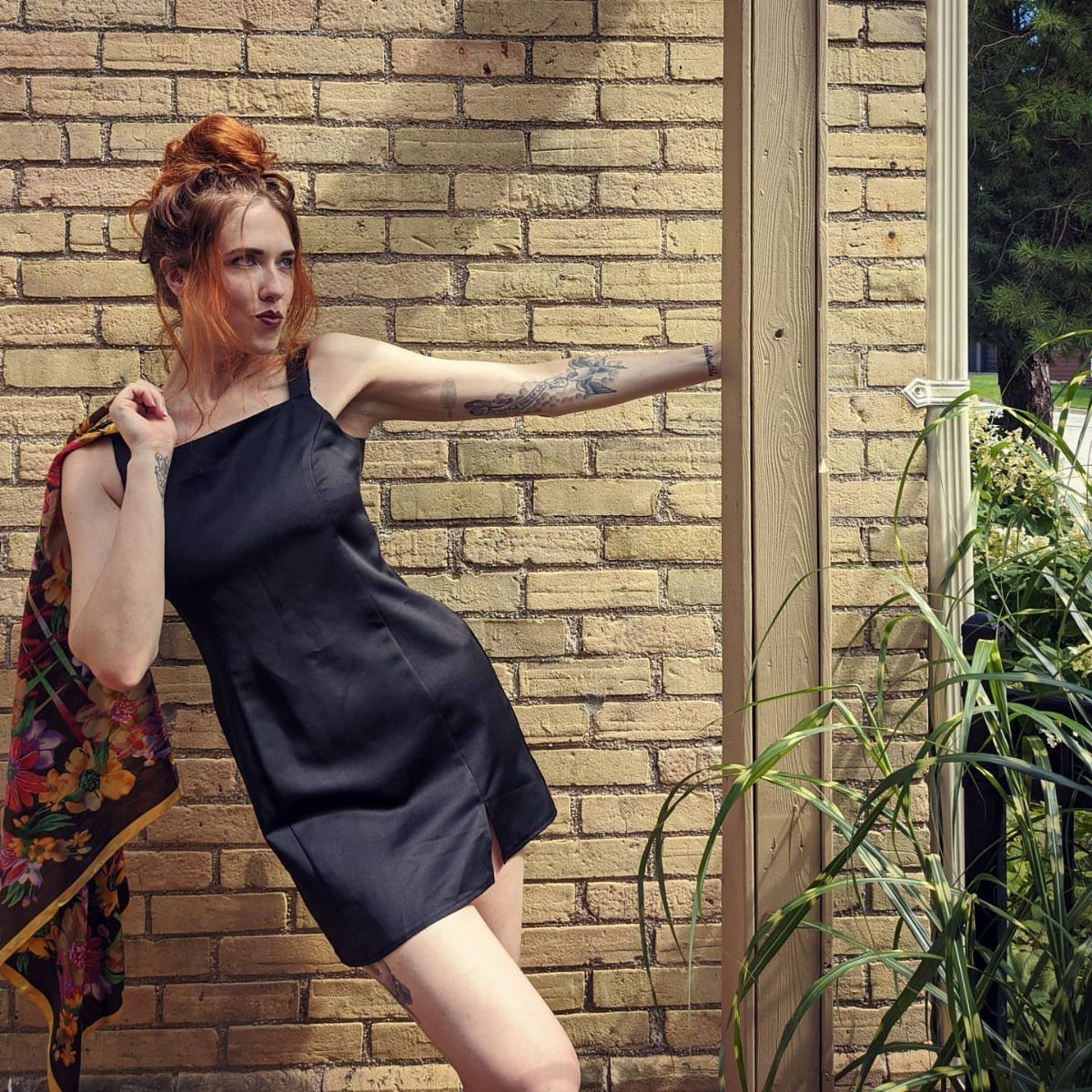 Woman with red hair wearing a black satin slip dress against a brick wall.