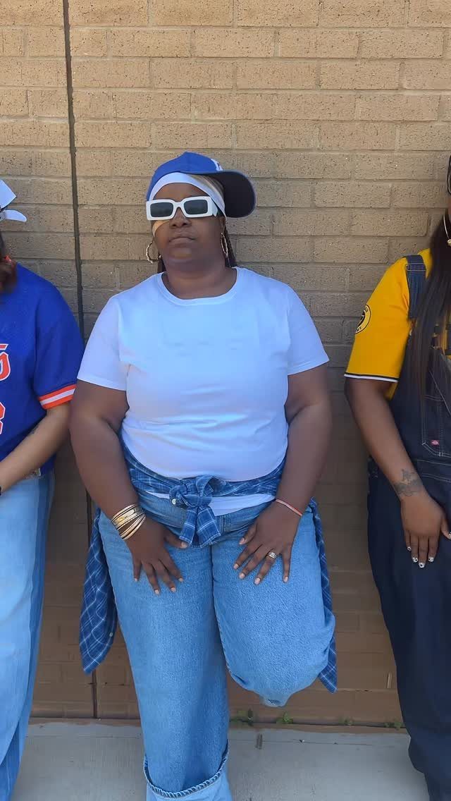 Woman wearing a white t-shirt, blue jeans, and a baseball cap against a brick wall.