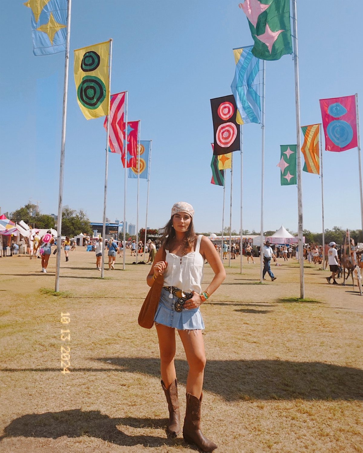 A woman in a white tank top and denim skirt posing in a festival field with colorful flags.