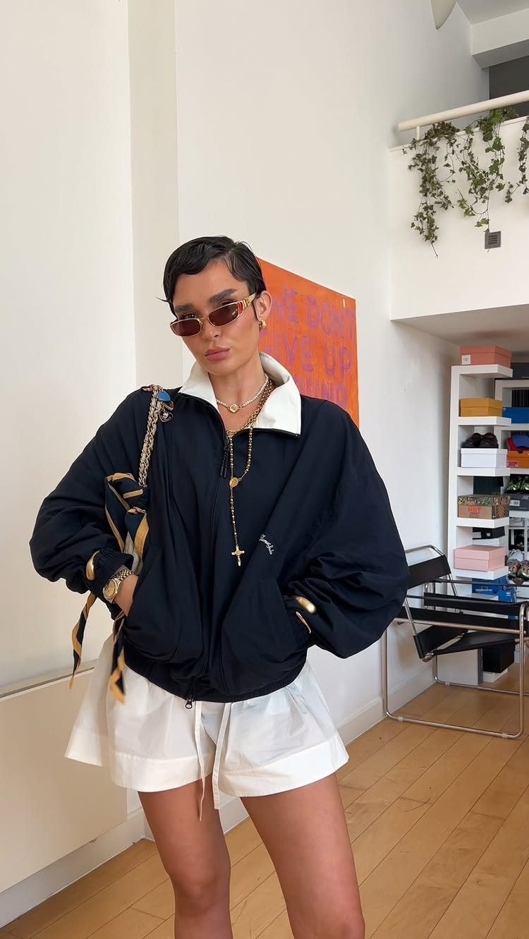 A woman wearing a black jacket, white shorts, and multiple layered gold necklaces.