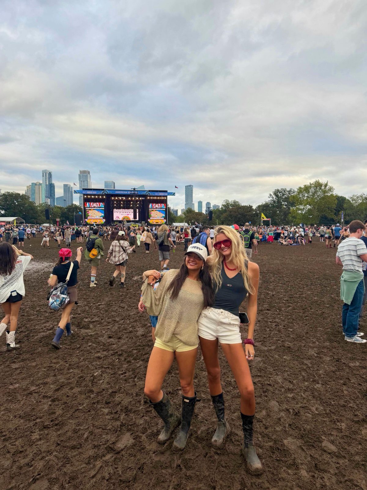 Two women in festival clothing and rubber boots posing in a muddy field near a stage.