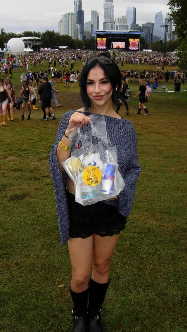 A smiling woman in a blue sweater and black skirt holding a bag at a music festival.