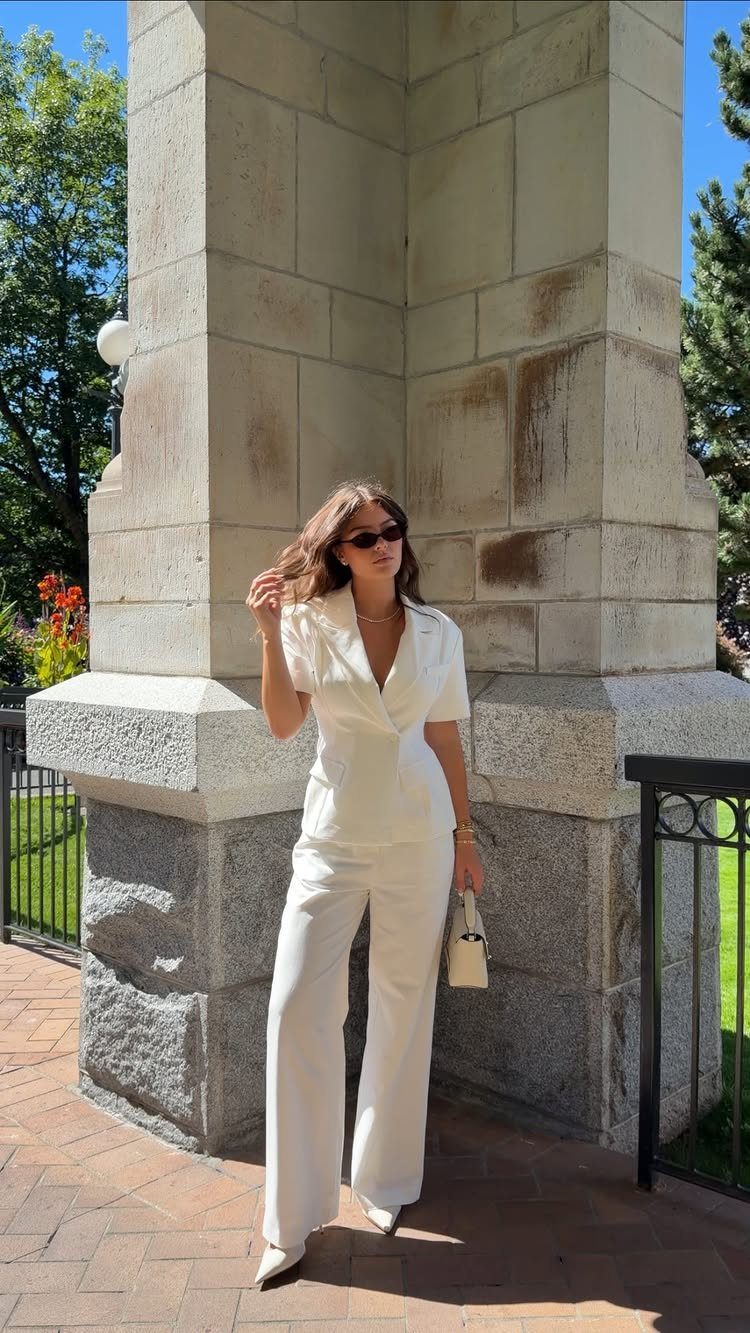 Woman in a white vest and wide-leg trousers standing by a stone pillar outdoors.