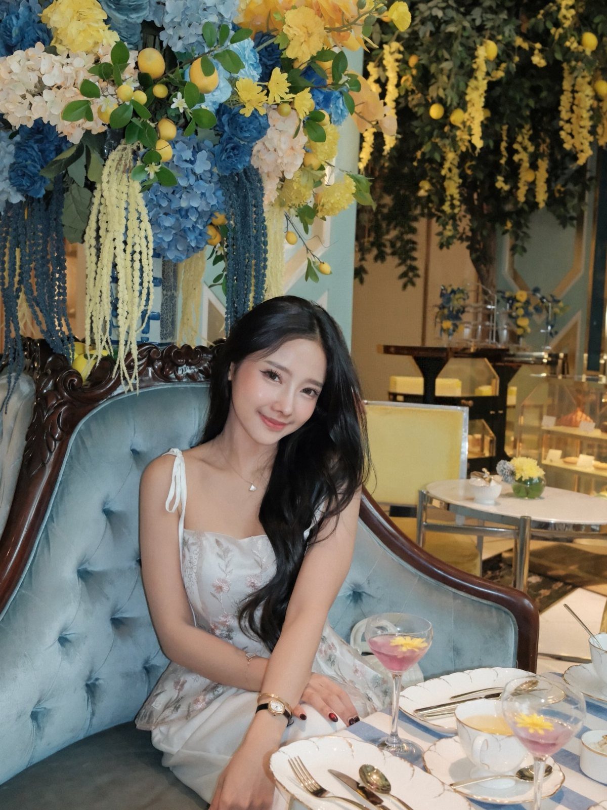 A young woman in a floral dress sits on a blue velvet sofa near large floral arrangements.