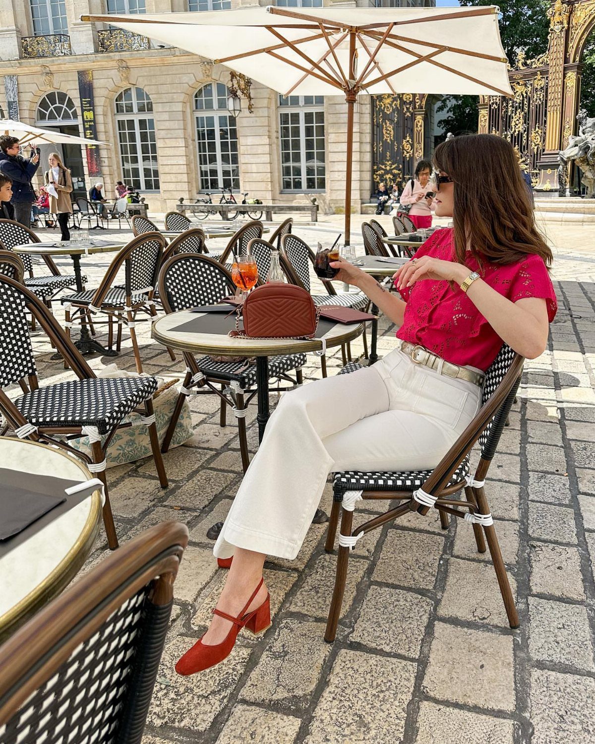 Woman in a pink blouse and white trousers sitting at an outdoor cafe table.