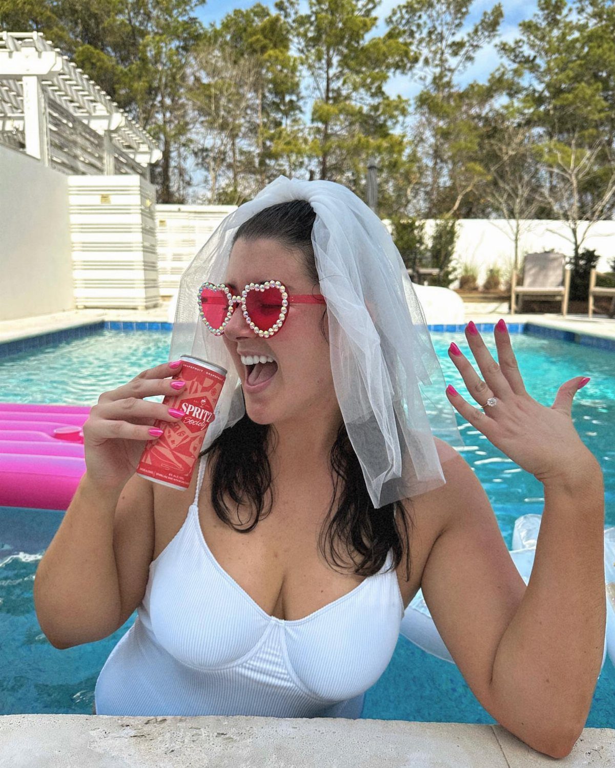 A woman wearing a white swimsuit and bridal veil smiles while holding a soda in a pool.