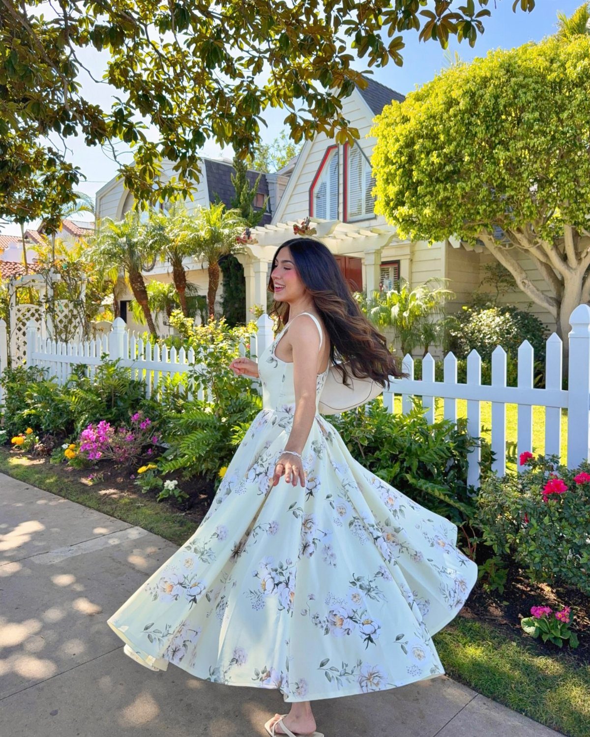 A woman twirling in a light yellow floral sundress next to a white picket fence.