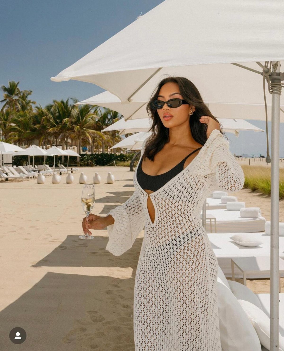A woman in a black bikini and white crochet cover-up holding champagne on a beach.