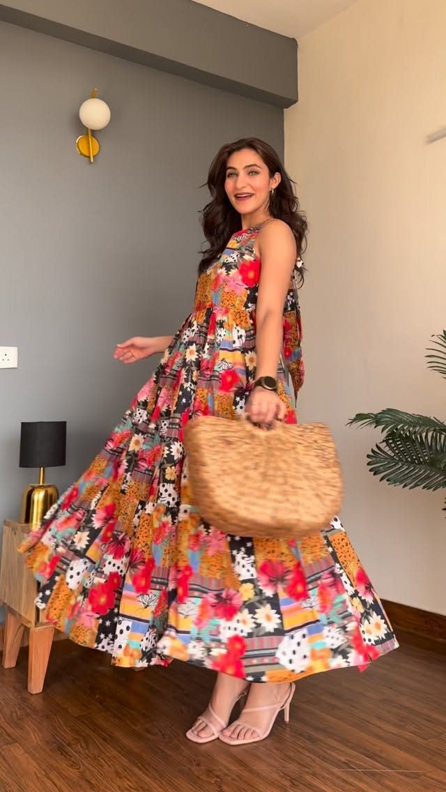 A woman in a colorful floral maxi dress holding a straw bag indoors.