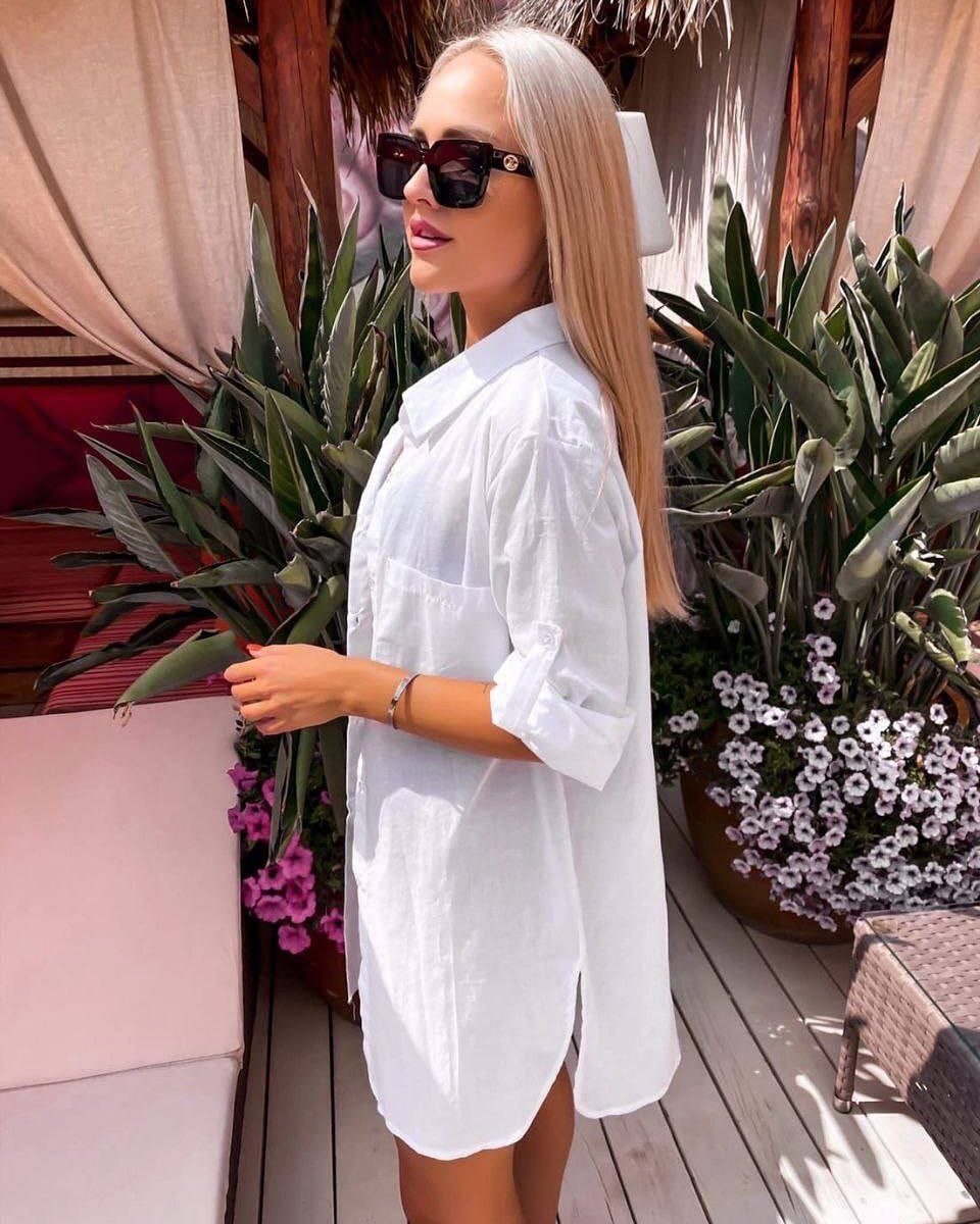 Blonde woman in sunglasses wearing a white poplin shirt dress standing near tropical plants.