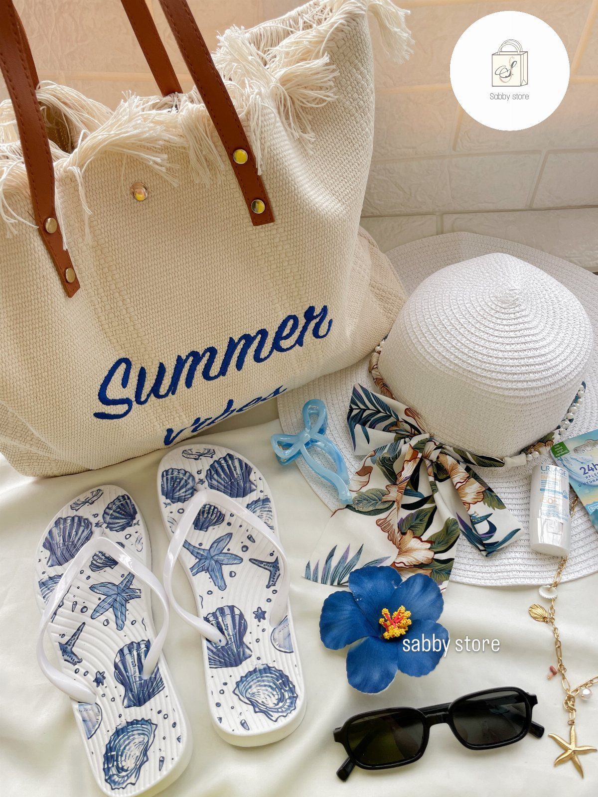 Beach accessories including a tote bag, sun hat, flip-flops, sunglasses, and starfish jewelry on white fabric.