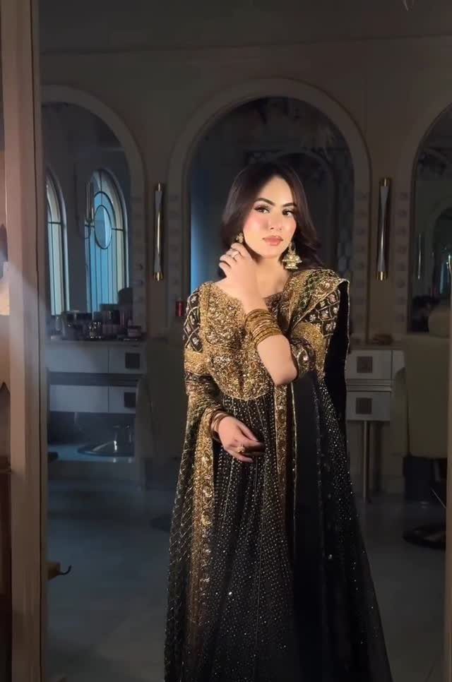 Woman in a black dress with intricate gold embroidery and gold jewelry posing indoors.