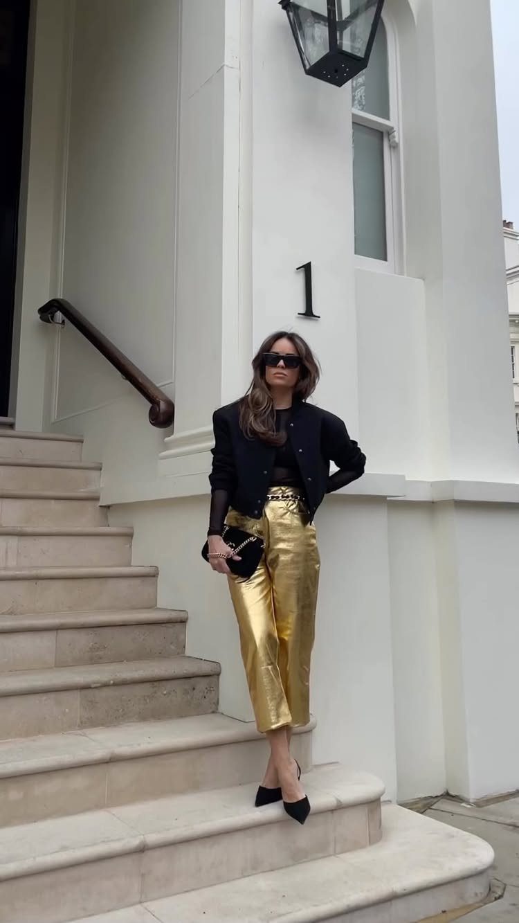 Woman posing on white stairs wearing a black jacket and metallic gold trousers.