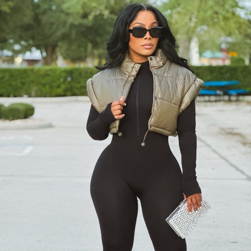 Woman in a black jumpsuit and metallic gold puffer vest posing outdoors with sunglasses.