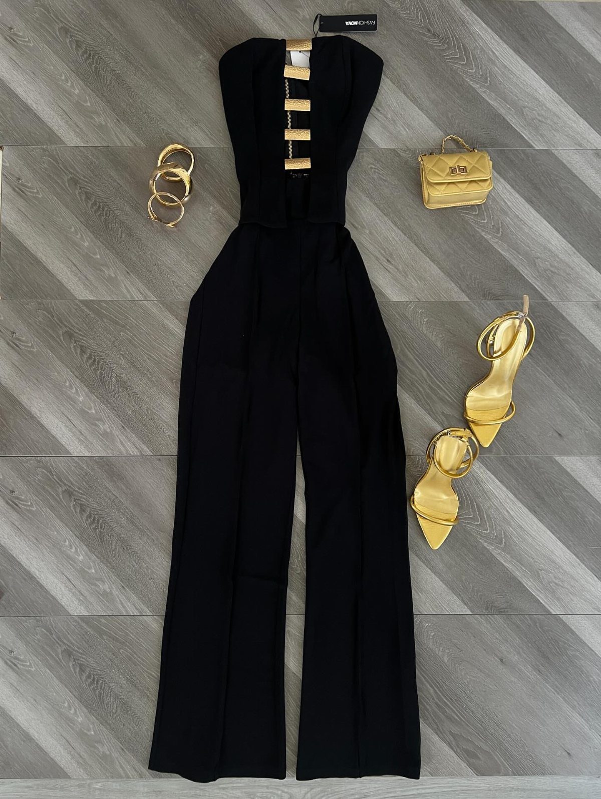 Black jumpsuit with gold accents laid flat with gold accessories and high heels on grey flooring.