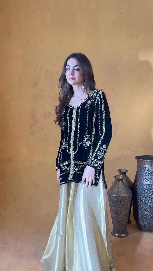Woman wearing a black embroidered tunic and a long gold metallic skirt standing against a gold background.