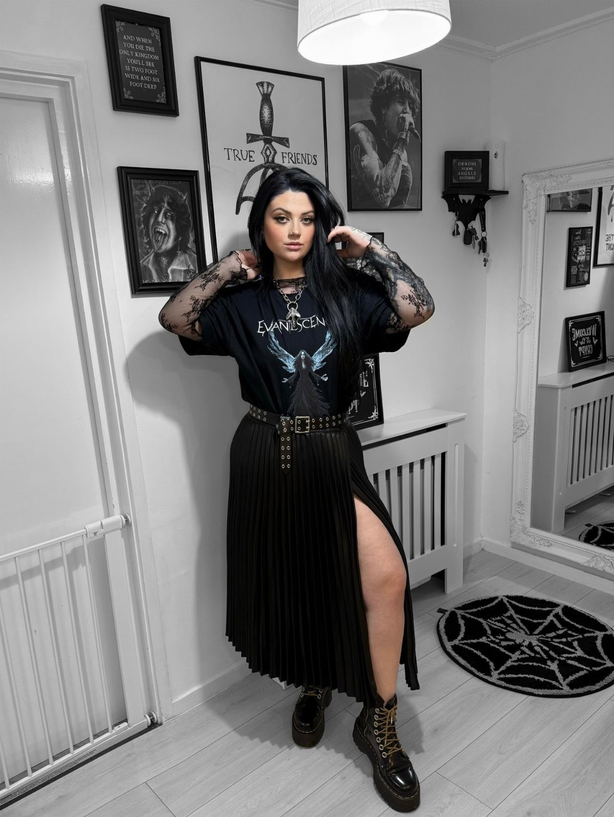 Woman posing in a black graphic t-shirt, pleated skirt, and lace-up boots in a decorated room.