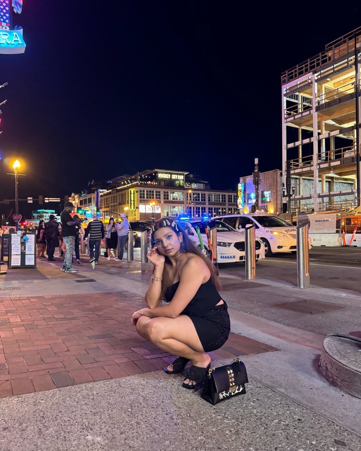 A woman in a black dress and platform sandals crouches on a city sidewalk at night.