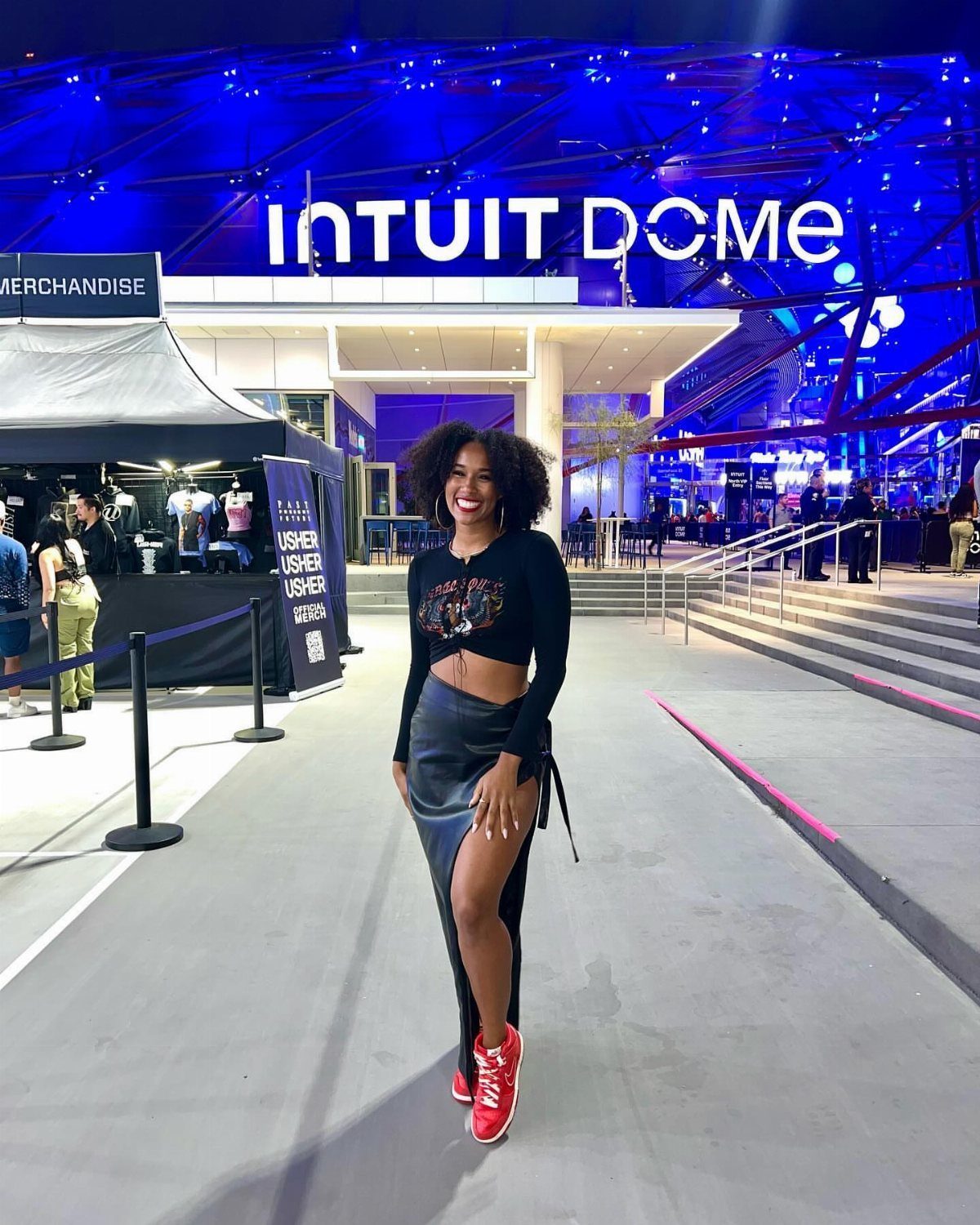 A smiling woman in a black concert outfit wearing bright red sneakers outdoors.