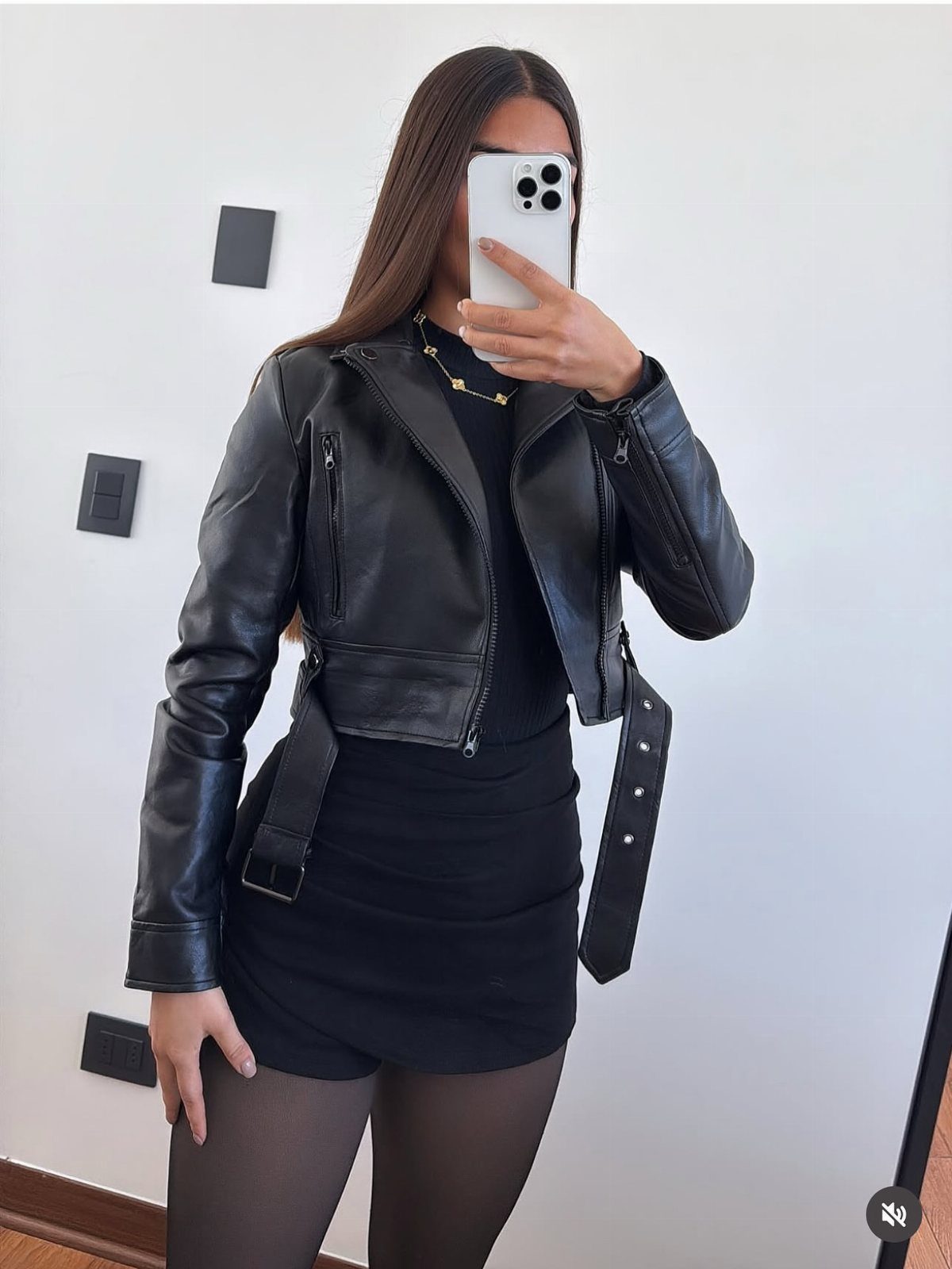 Woman in a black leather biker jacket, black mini skirt, and sheer tights posing for a selfie.