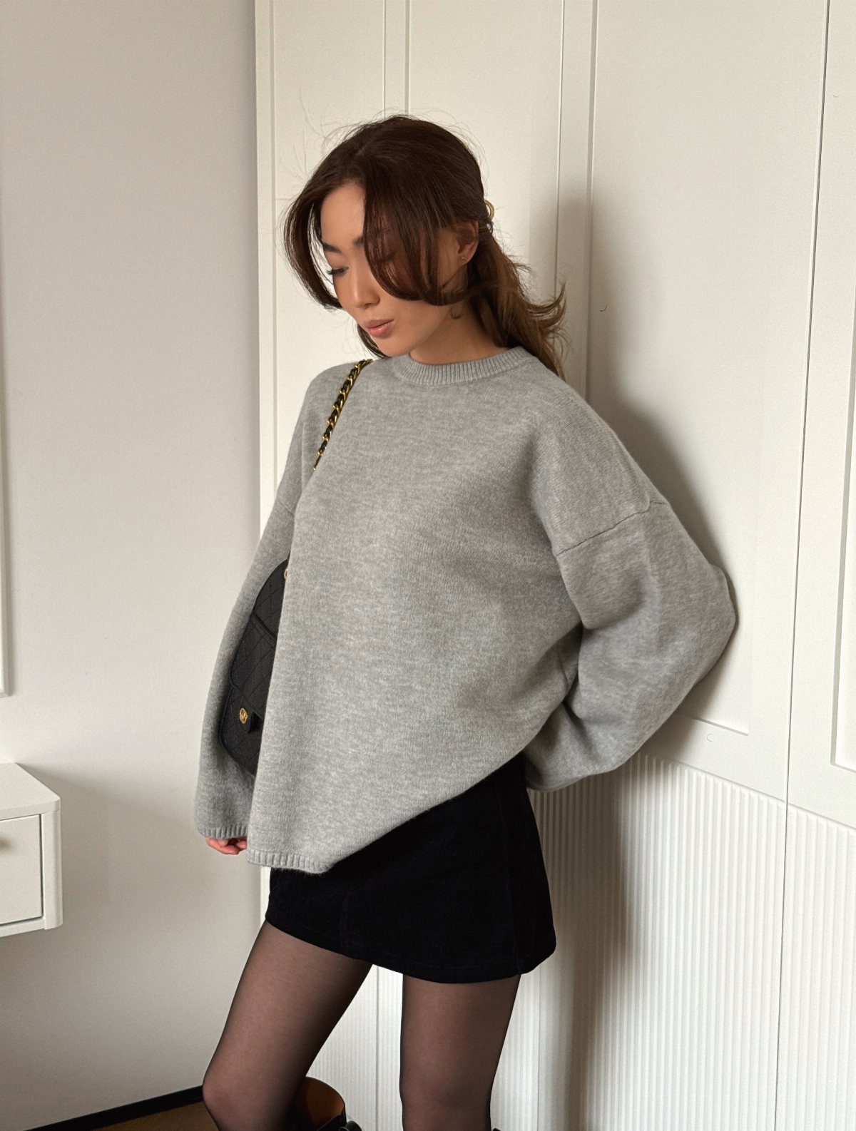Woman in a grey oversized sweater, black mini skirt, and sheer black tights standing by a wall.