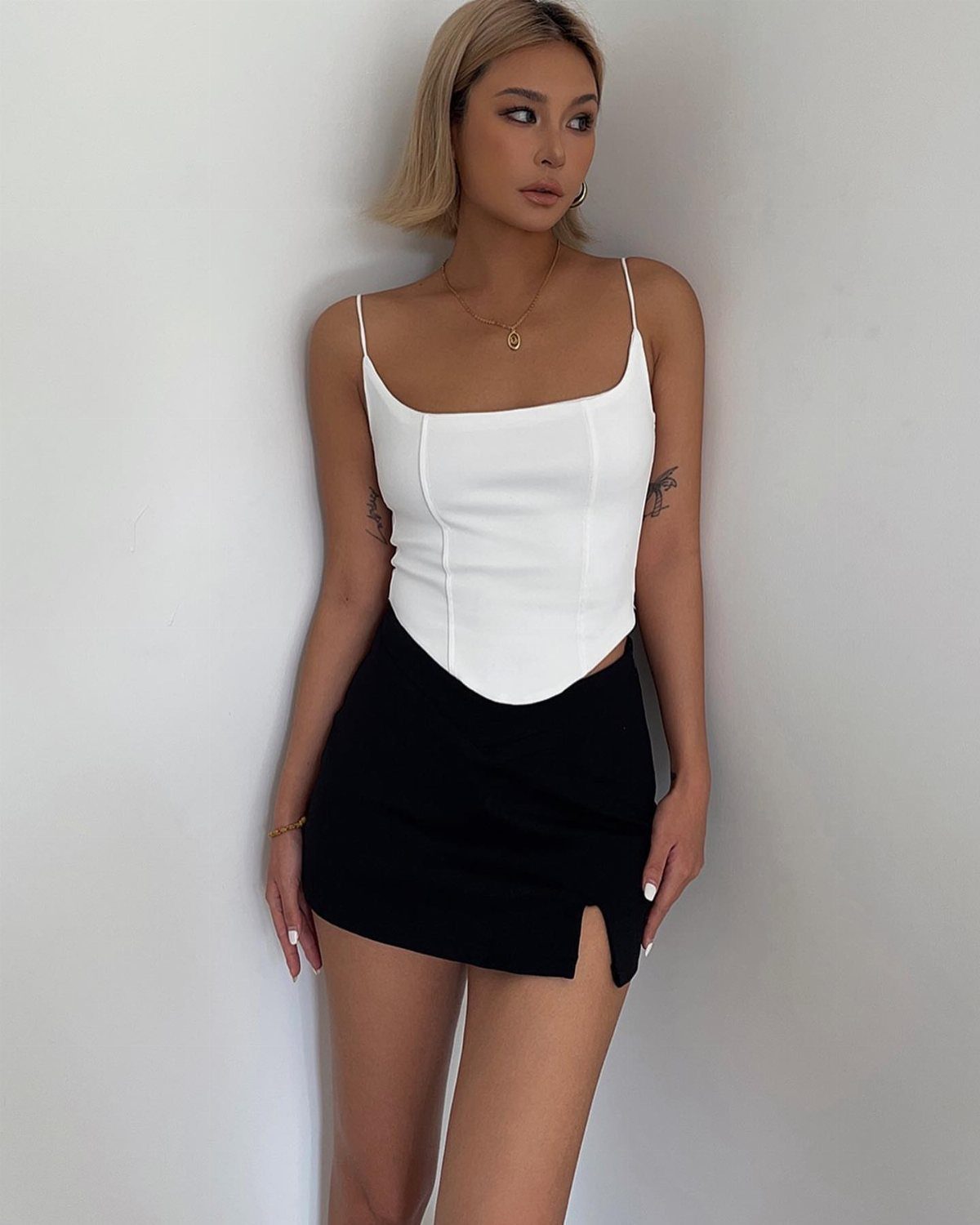 A blonde woman posing in a white camisole and black mini skirt against a white wall.