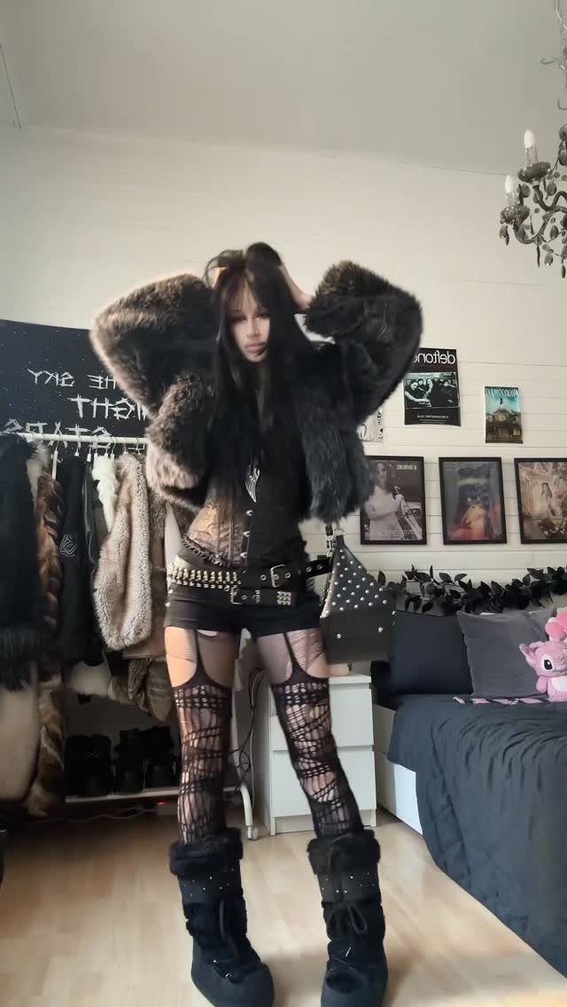 Woman posing in black rave clothing with a large faux fur jacket and fishnet stockings.