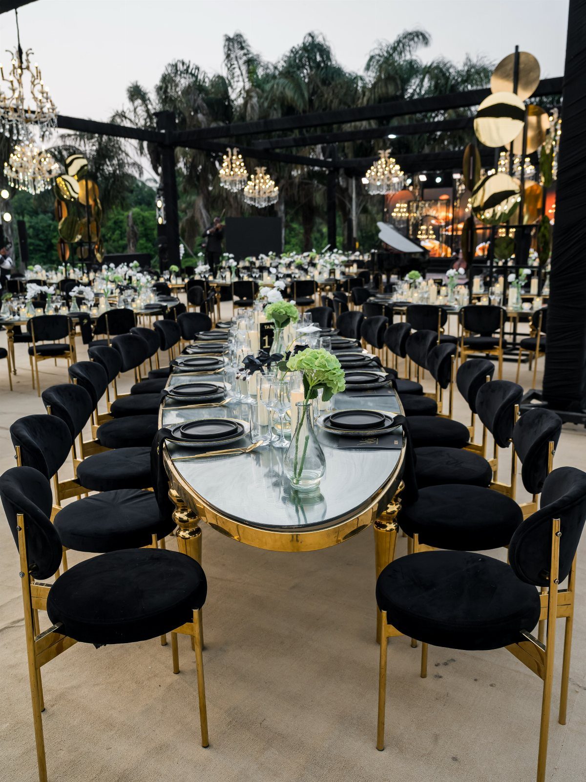 Black velvet and gold chairs arranged around long glass-topped banquet tables at an outdoor event.