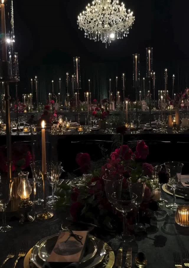 Elegant formal dinner tables set with tall candles, red flowers, and a crystal chandelier in dark lighting.