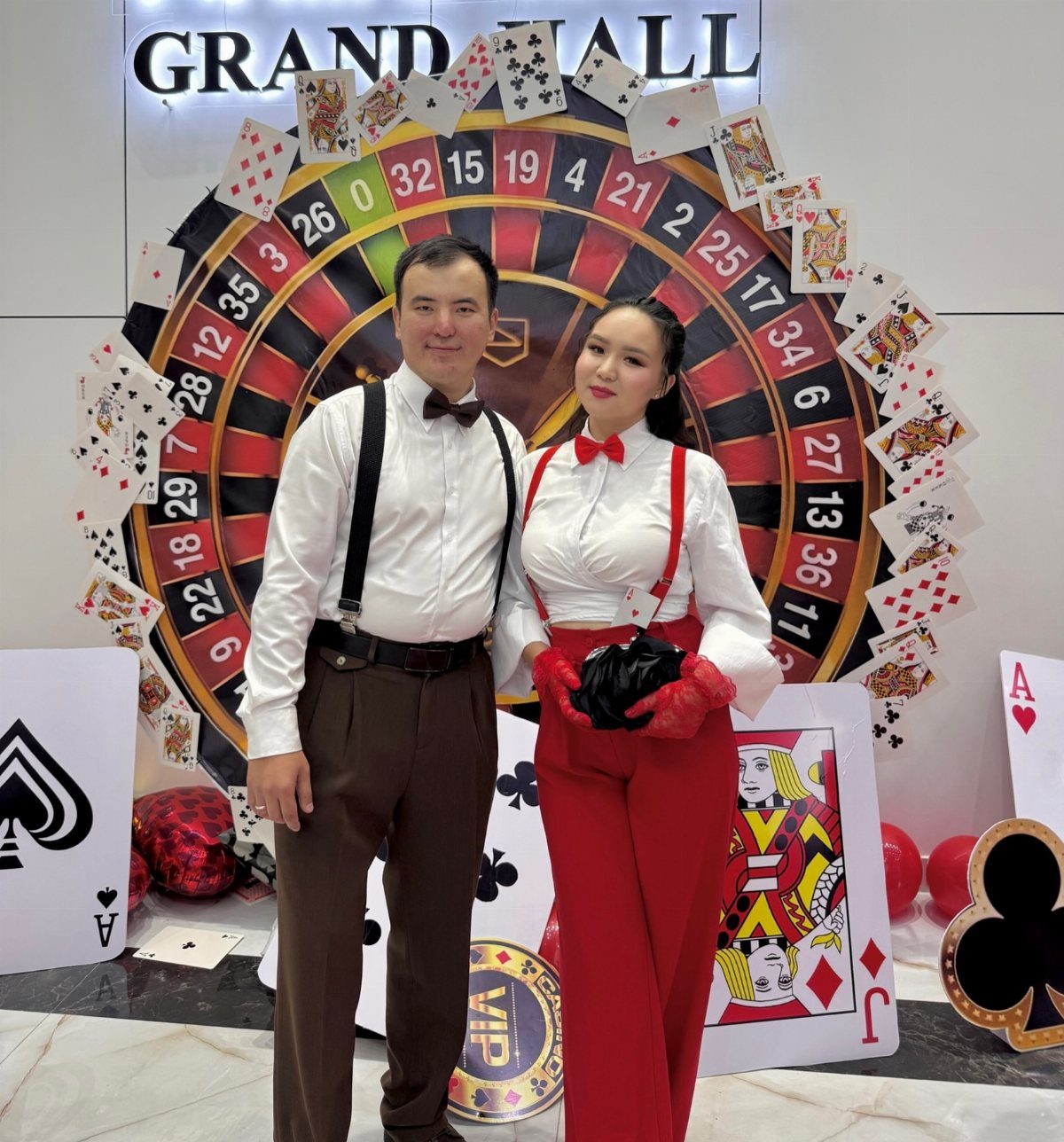 A man and woman posing in themed outfits in front of a large roulette wheel backdrop.