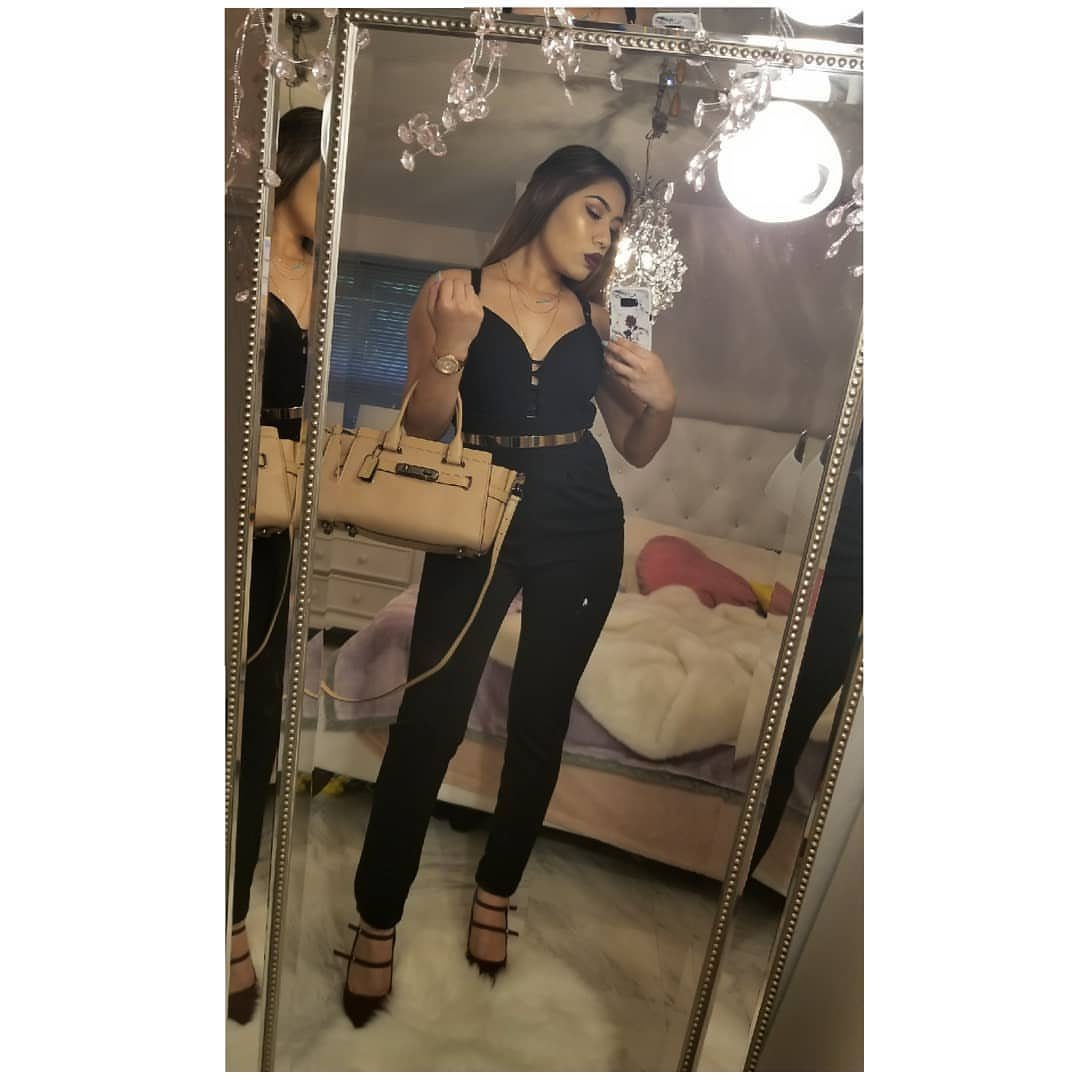 Woman in a black jumpsuit with a gold belt posing for a mirror selfie.