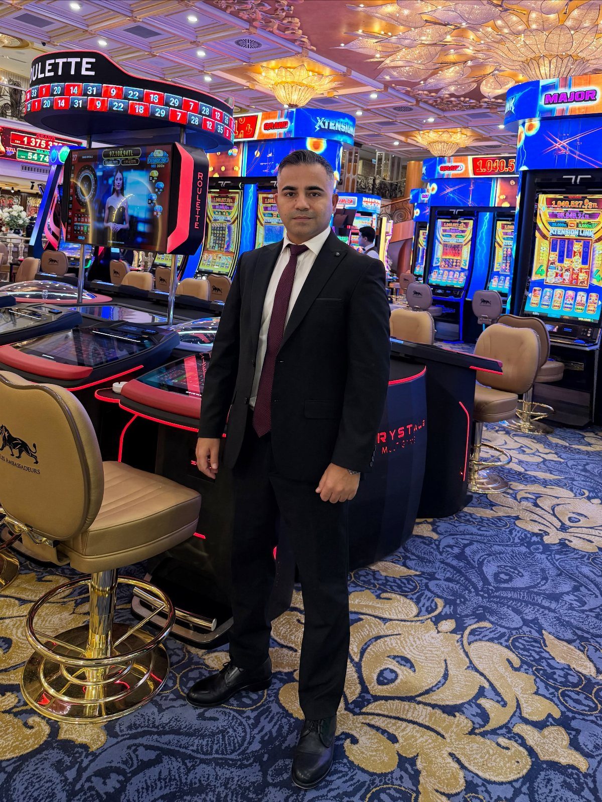 A man wearing a black suit and red tie standing in a casino setting.