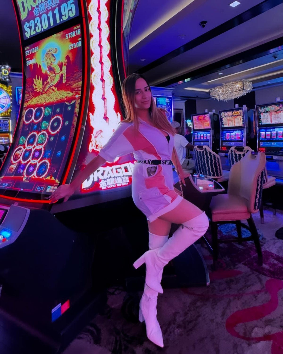 Woman in a white mini dress and thigh-high boots posing by a slot machine.