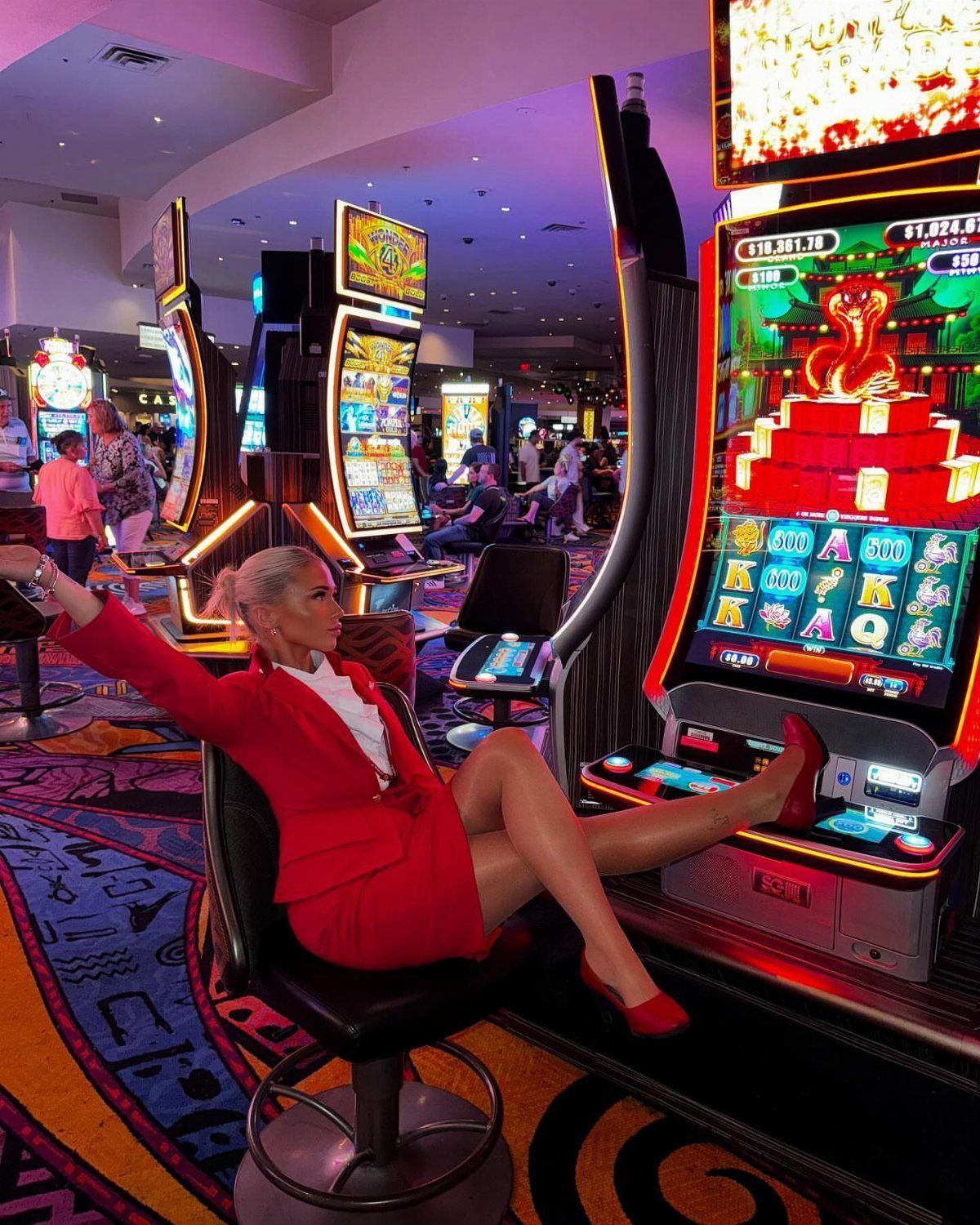 Woman in a red suit and heels sitting at a slot machine in a casino.