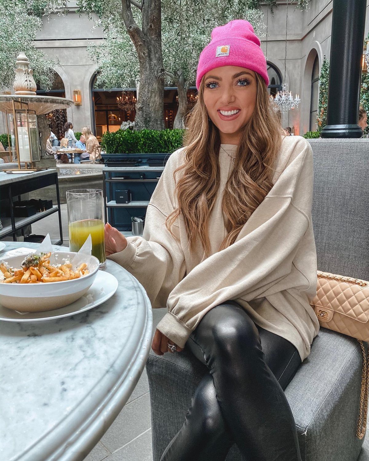 A smiling woman wearing a bright pink beanie and a beige sweater at an outdoor cafe.