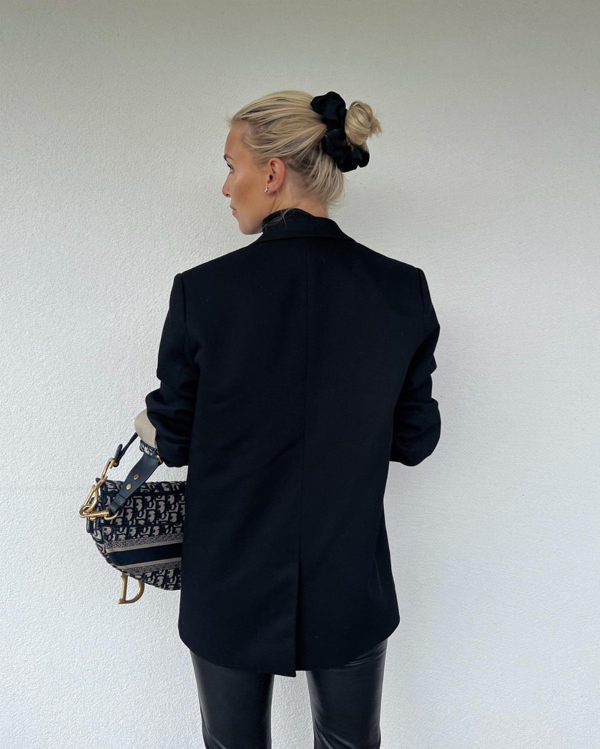 A woman wearing a black blazer and black leather pants standing against a white wall.