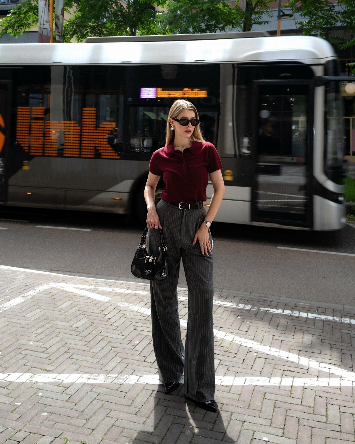 Woman in a burgundy polo shirt and grey pinstriped trousers standing near a city bus.