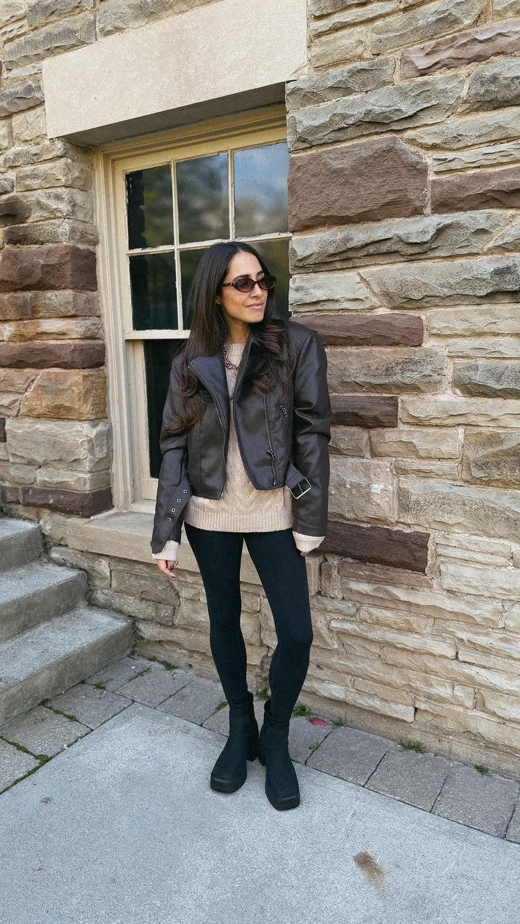 Woman wearing a leather jacket, beige sweater, black leggings, and black Chelsea boots standing by a stone wall.
