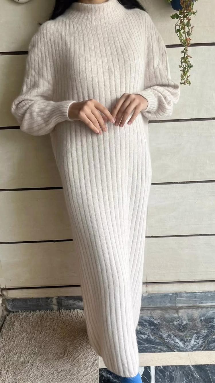 A person wearing a long, cream-colored ribbed knit sweater dress standing against a wall.