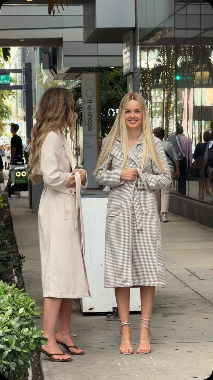 Two women standing on a city sidewalk wearing long, belted trench coats and stylish footwear.